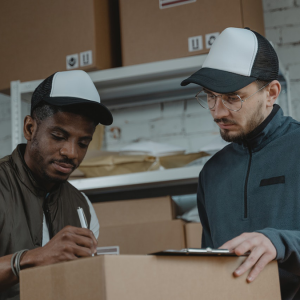 Man filling out a form while another man holds a box in a warehouse